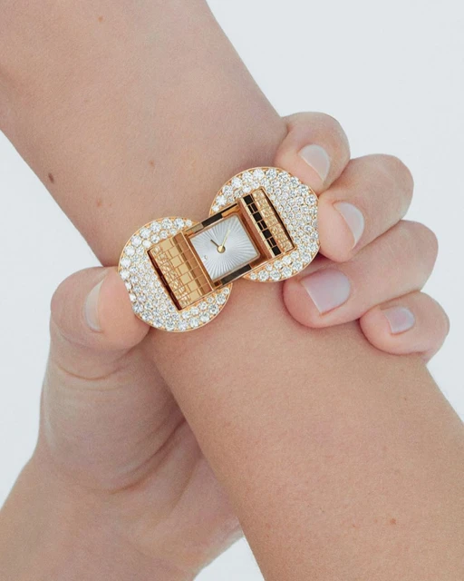 Wrist shot of a person wearing a luxury gold watch with diamond embellishments. The jewelry is in focus against a plain background.