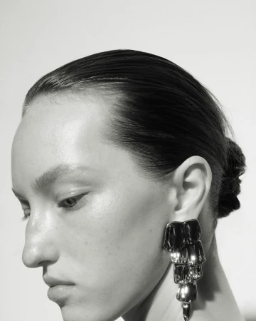 Close-up profile of a woman with slicked-back dark hair wearing a large metallic statement earring against a white background.