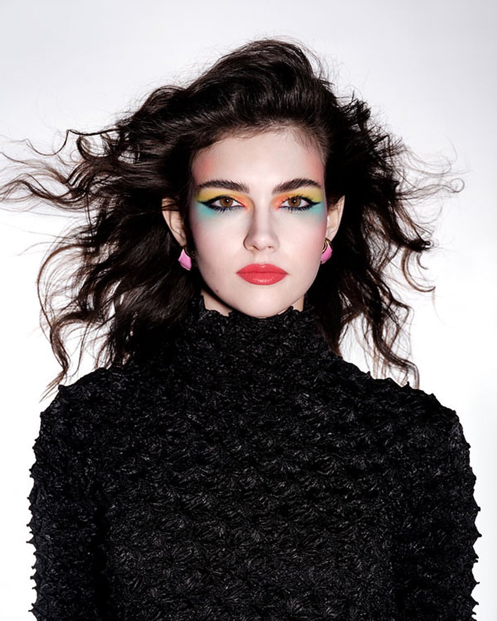Portrait of a woman with dark, wavy hair, rainbow eye makeup, and a black high-neck top, set against a bright white backdrop in a studio shot.