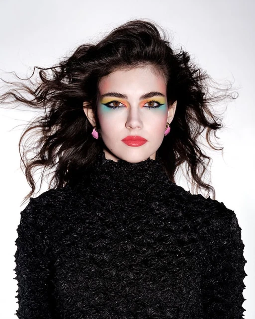 Portrait of a woman with dark, wavy hair, rainbow eye makeup, and a black high-neck top, set against a bright white backdrop in a studio shot.