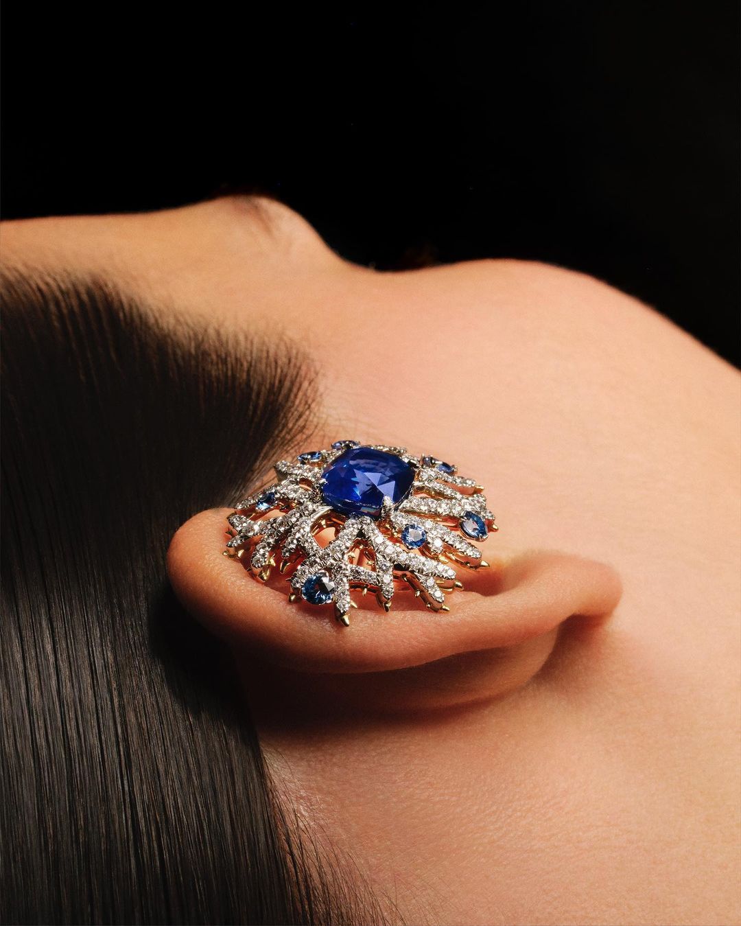 Closeup of a woman's ear adorned with an elaborate gold and blue gemstone earring in a studio setting against a dark backdrop.