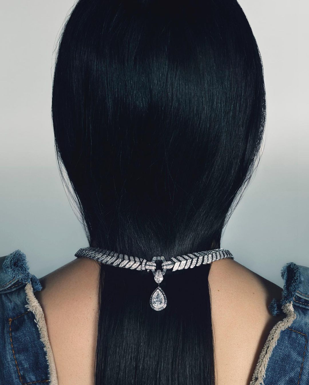 Close-up of a woman's back with black hair wearing a white jeweled necklace and a denim jacket against a white background.