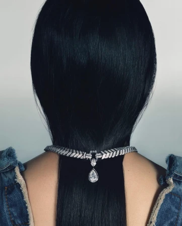 Close-up portrait from behind of a person with long straight black hair wearing a diamond pendant necklace over denim clothing.