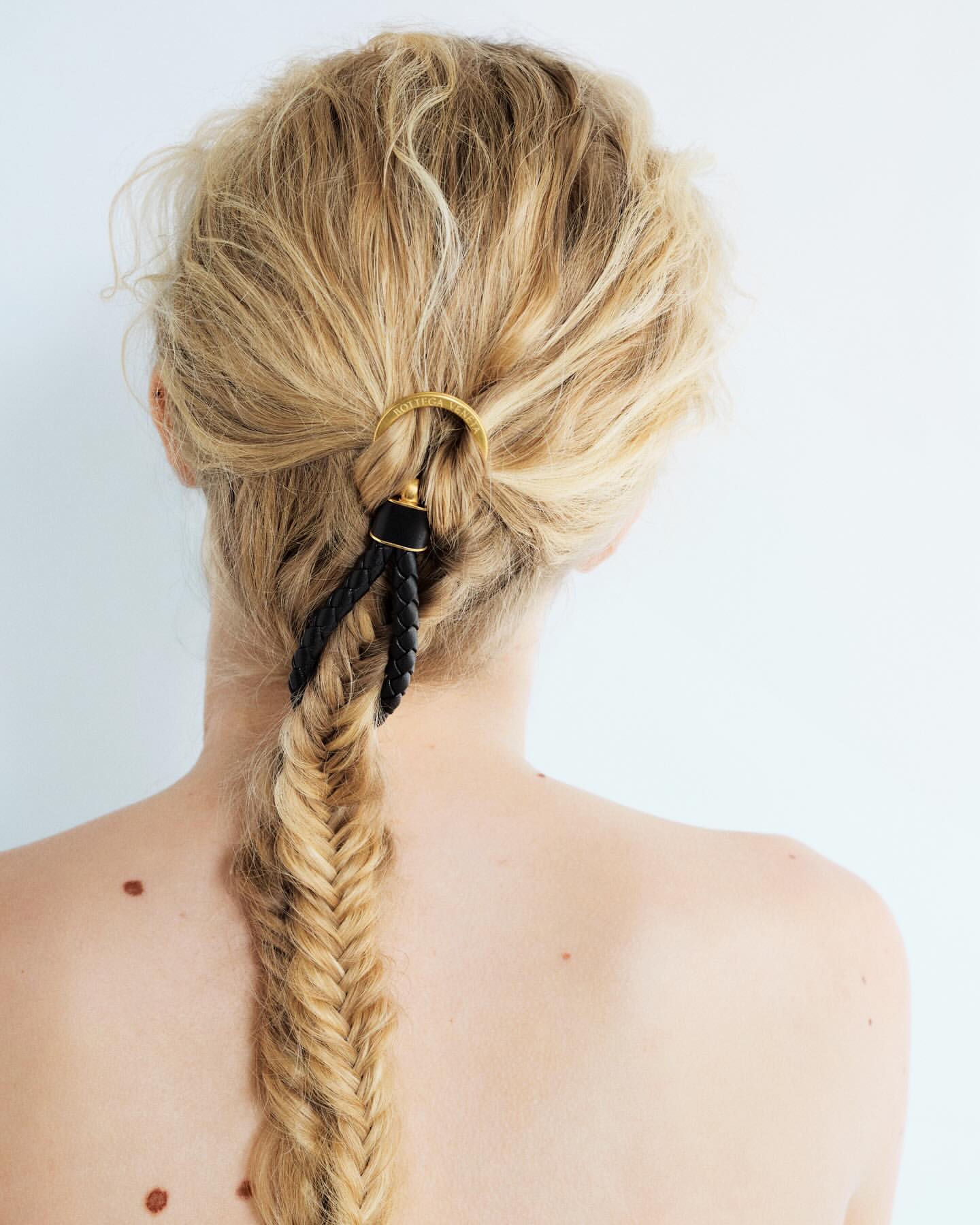 Woman's blonde hair styled in a long braid with a gold and black decorative hair accessory, back view against a white background.