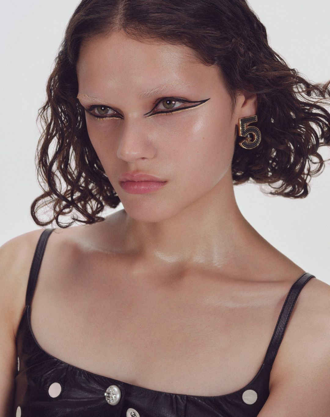 Portrait of a young woman with bold black eyeliner and a black top, accessorized with a prominent gold earring shaped like the number '5'.