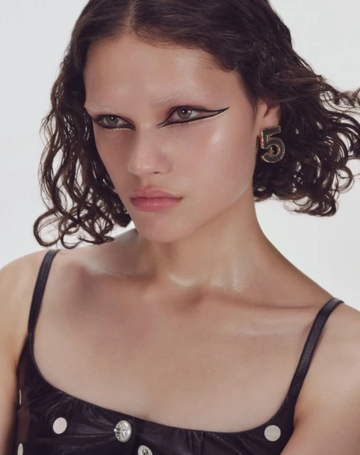 Portrait of a young woman with bold black eyeliner and a black top, accessorized with a prominent gold earring shaped like the number '5'.