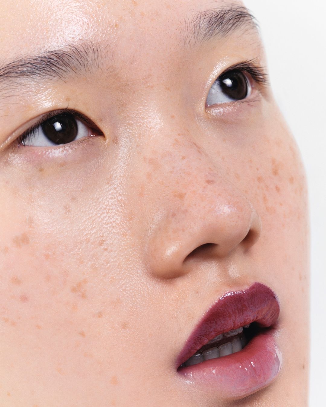 Close-up portrait of an Asian female face with freckles, red lipstick, and glossy skin against a bright white background, shot with eye-level angle.