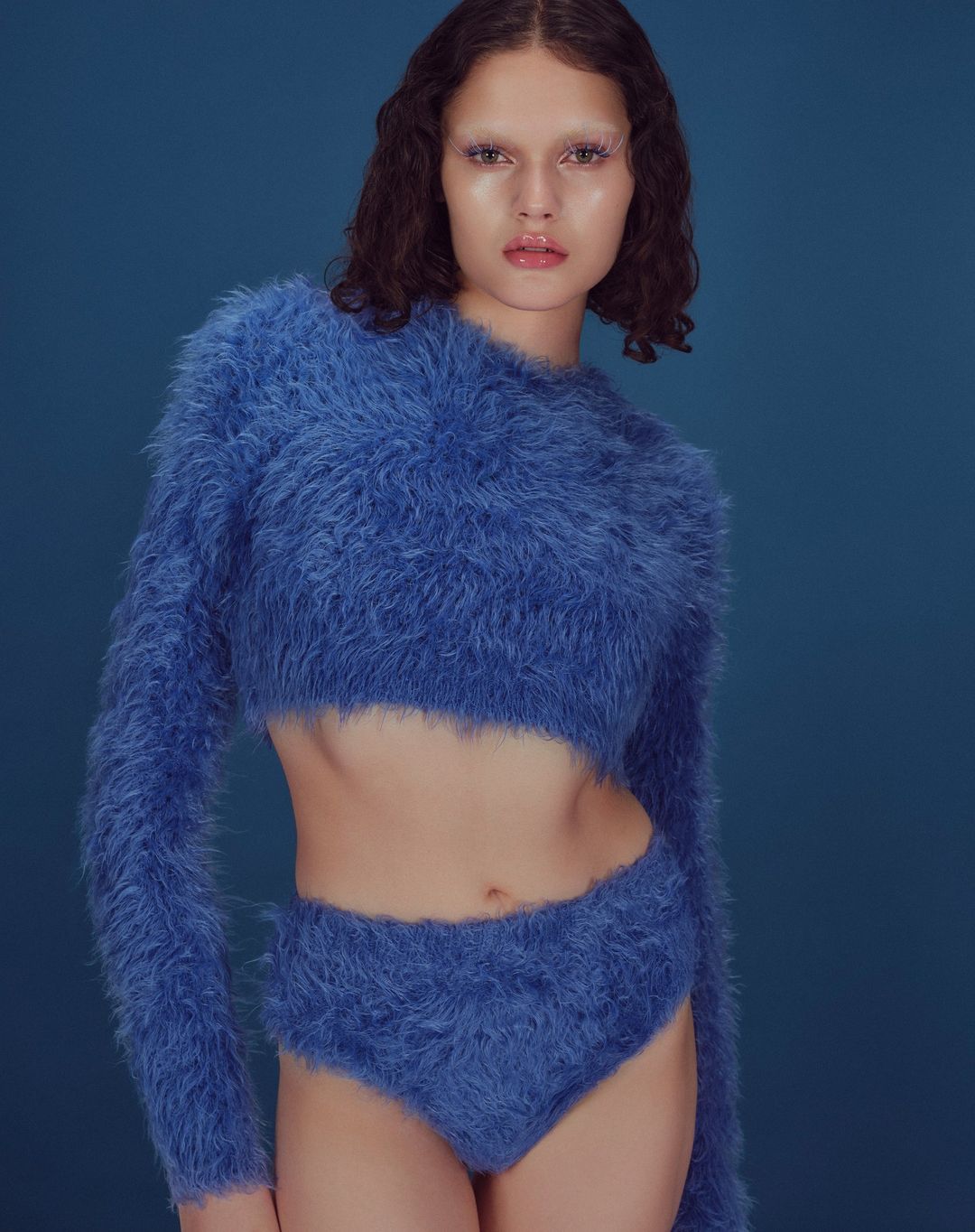 Woman in a blue fuzzy sweater and high-waisted bottoms poses against a solid blue backdrop, presenting a fashion-forward aesthetic.