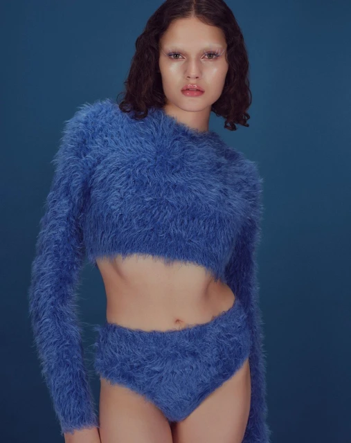 Woman in a blue fuzzy sweater and high-waisted bottoms poses against a solid blue backdrop, presenting a fashion-forward aesthetic.