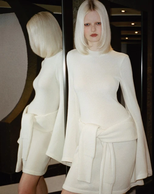 A fair-skinned woman with platinum blonde hair poses in a white dress with bell sleeves, with her reflection visible in a dark mirror.
