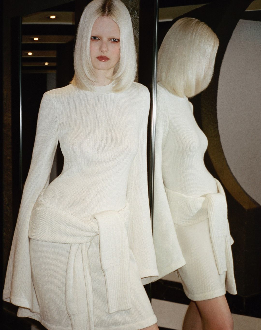 Blonde woman in a long-sleeve mini dress poses in front of a mirror, reflecting her image, creating a visually striking, fashion-forward scene.