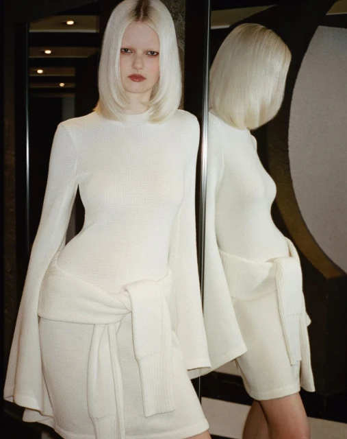 Blonde woman in a long-sleeve mini dress poses in front of a mirror, reflecting her image, creating a visually striking, fashion-forward scene.