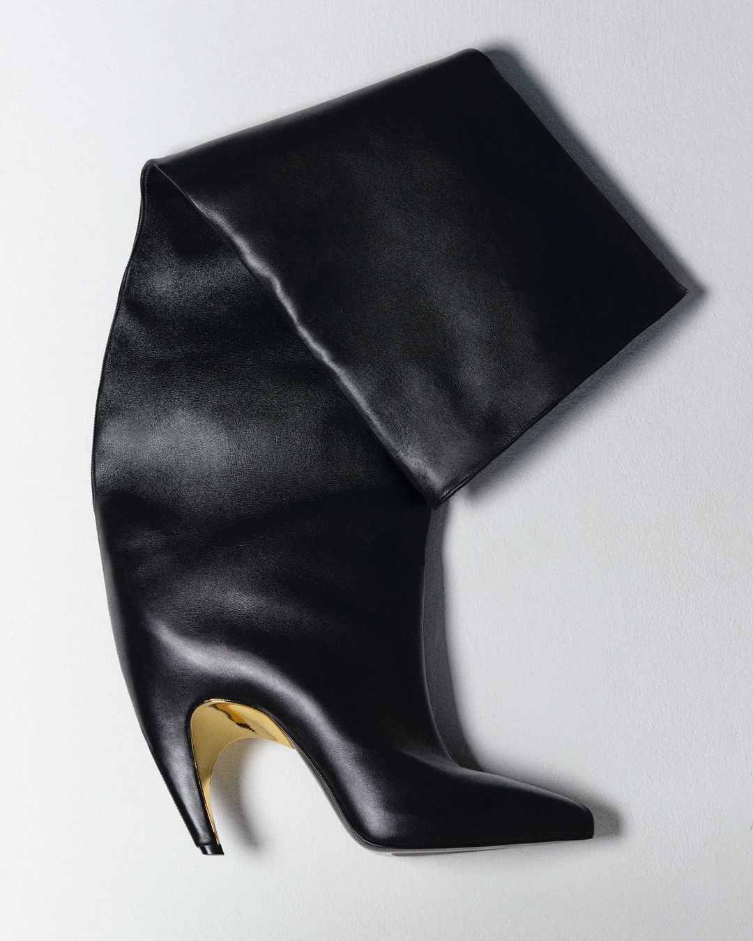 Studio shot of a sleek black, over-the-knee, high-heeled boot with a metallic gold heel, set against a clean, light background.