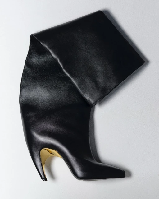 Studio shot of a sleek black, over-the-knee, high-heeled boot with a metallic gold heel, set against a clean, light background.