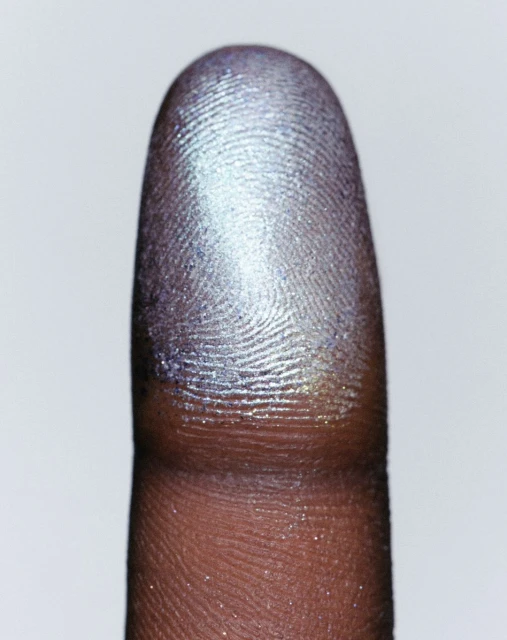 Macro photograph showcasing a close-up of a human fingerprint with iridescent cosmetic shimmer on the fingertip against a plain white backdrop.