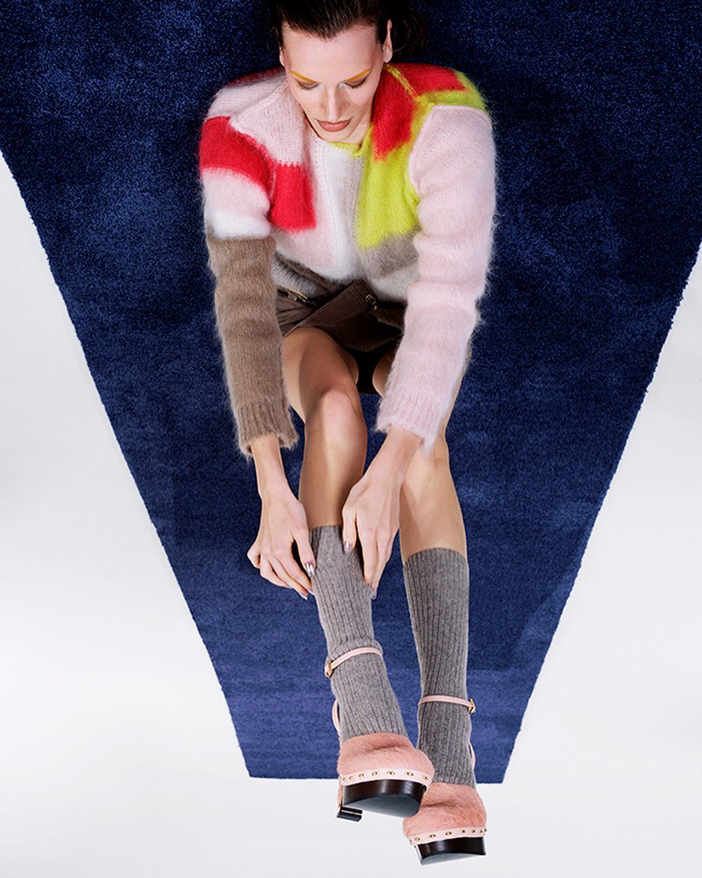Overhead shot of a stylish woman with a multicolored sweater, mini skirt, socks, and platform heels, posing on a blue and white carpet.