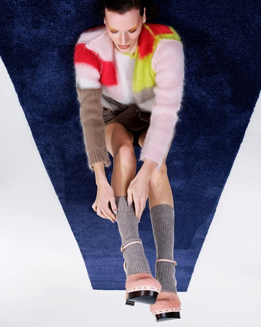 Overhead shot of a stylish woman with a multicolored sweater, mini skirt, socks, and platform heels, posing on a blue and white carpet.