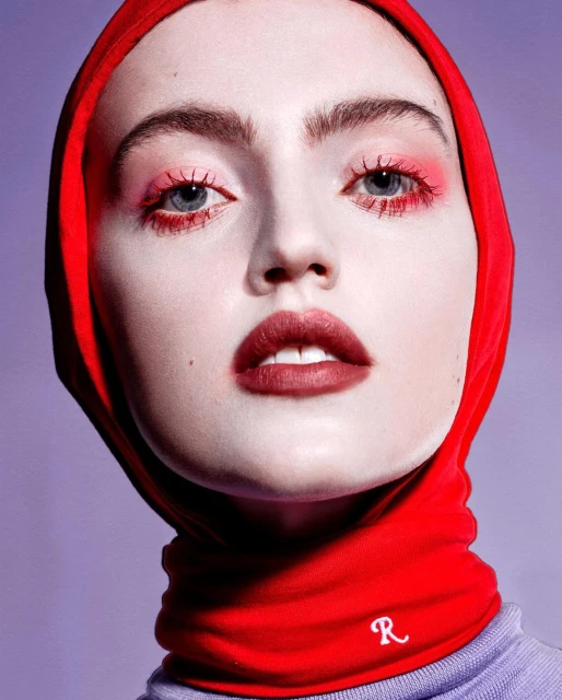 Headshot of a fair-skinned woman with red eye makeup and lipstick, wearing a red headscarf and lilac sweater, on a light purple background.