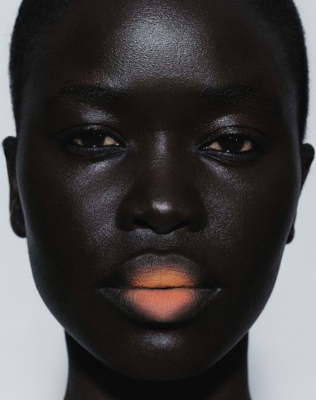 Close-up portrait of a Black woman with dramatic lighting highlighting her skin's texture, with peach ombre lipstick against a white background.
