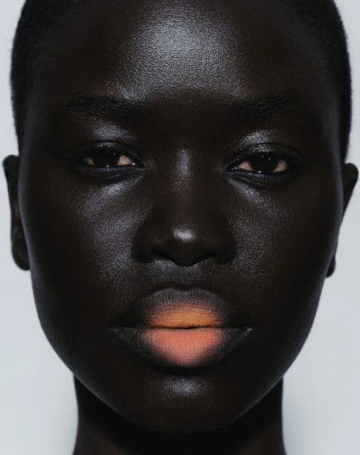 Close-up portrait of a Black woman with dramatic lighting highlighting her skin's texture, with peach ombre lipstick against a white background.