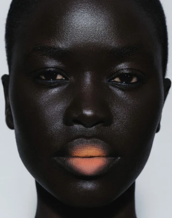 Close-up portrait of a Black woman with dramatic lighting highlighting her skin's texture, with peach ombre lipstick against a white background.