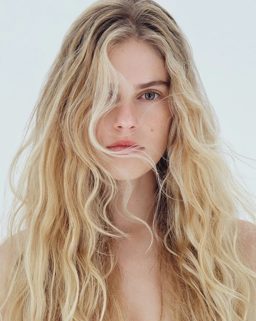 Portrait of a blond adult female with long, wavy hair partially covering her face, looking at the camera against a bright white backdrop.