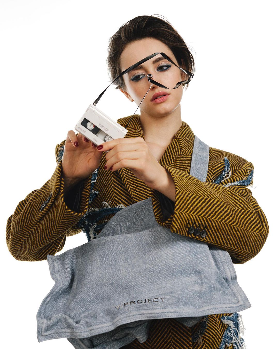 Woman in a herringbone jacket holds a vintage cassette player with glasses hanging on it, plus a denim bag in a bright photo studio.