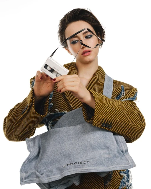 Woman in a herringbone jacket holds a vintage cassette player with glasses hanging on it, plus a denim bag in a bright photo studio.