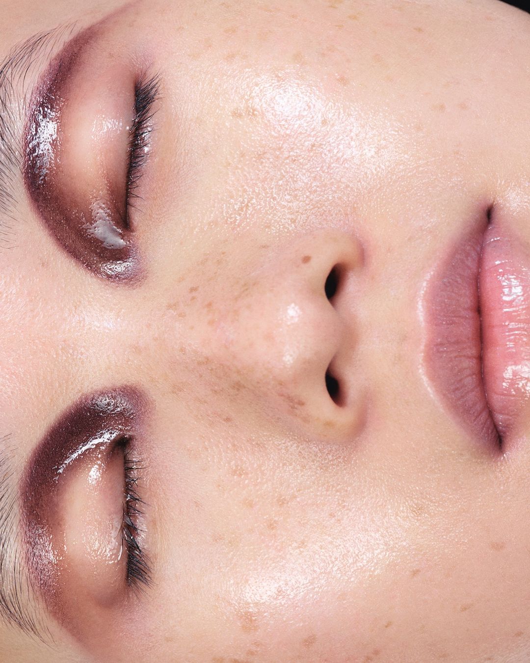 Extreme close-up portrait of a face with glossy burgundy eyeshadow, glossy lips, and visible freckles shot by Antoine And Charlie.