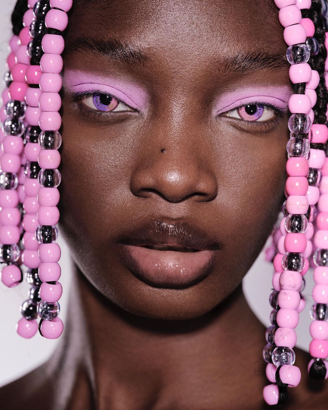 Close-up portrait of an African-American adult female with pink and clear beads in her hair and wearing lilac makeup.