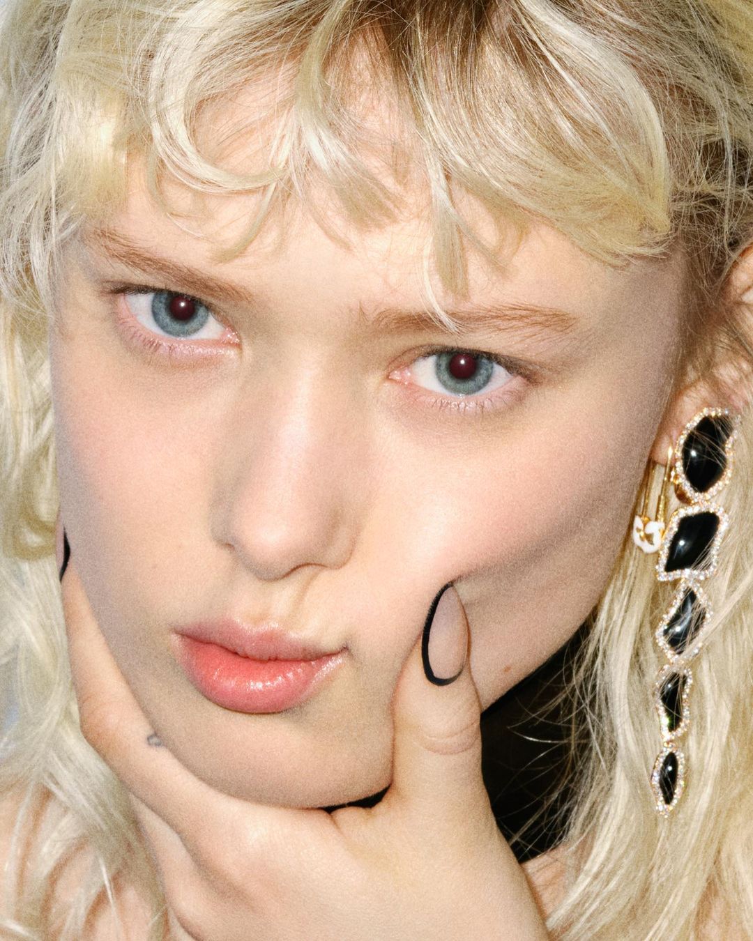 Close-up portrait of a young adult female with blonde wavy hair, light skin, light blue eyes, glossy lips, a black and gold earring, and manicured nails.