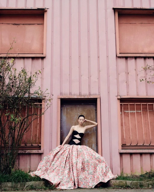 A model in a strapless black and floral gown poses in front of a light pink corrugated metal building with boarded-up windows and a door.