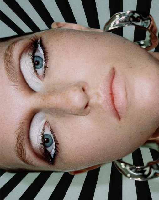 Close-up beauty shot of a fair-skinned young woman with silver hoop earrings against a high-contrast black-and-white striped backdrop.