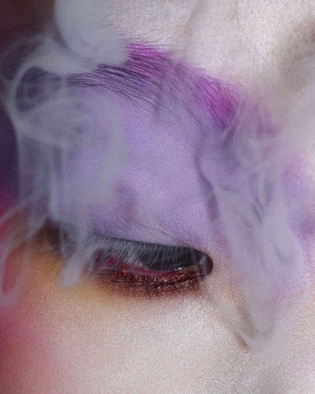 Close-up studio shot of an adult female face with closed eyes, purple makeup, and vapor-like smoke around her face.