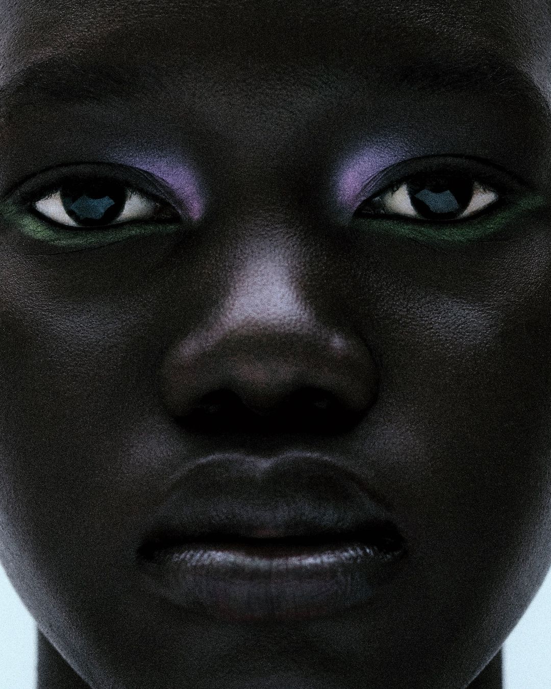 Close-up portrait of a dark-skinned person with colorful makeup on their eyes, set against a pale background in a beauty shot.
