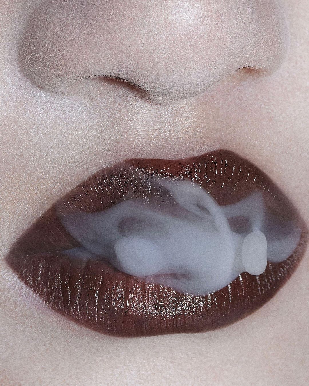 Close-up of a woman's face with dark red lipstick, exhaling a cloud of white smoke from her mouth in a beauty shot with smooth skin.