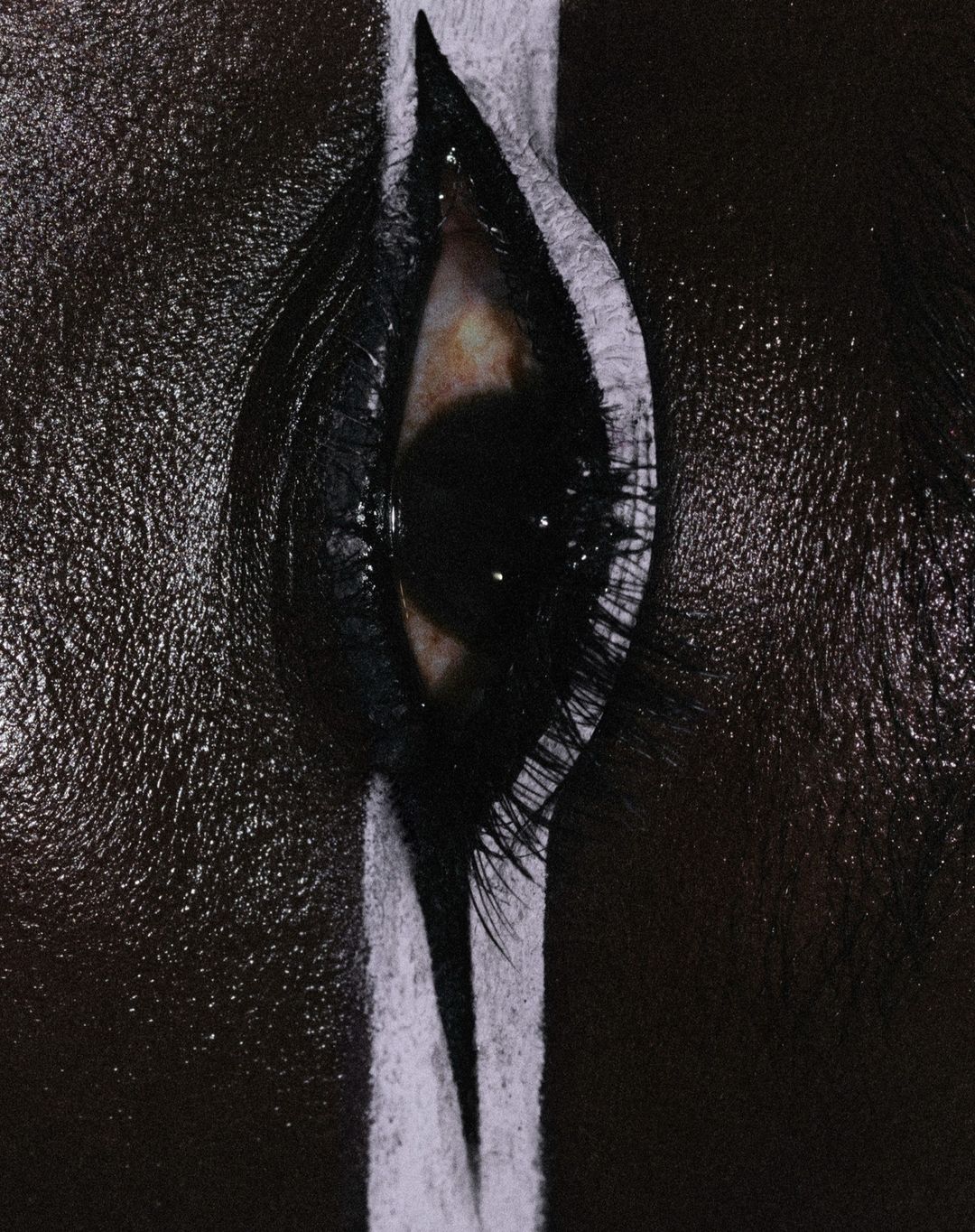 Close-up of a single eye with heavy makeup and a thick, white stripe running vertically over the lid and lashes, contrasted against dark skin.