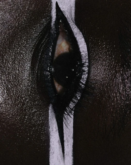 Close-up of a single eye with heavy makeup and a thick, white stripe running vertically over the lid and lashes, contrasted against dark skin.