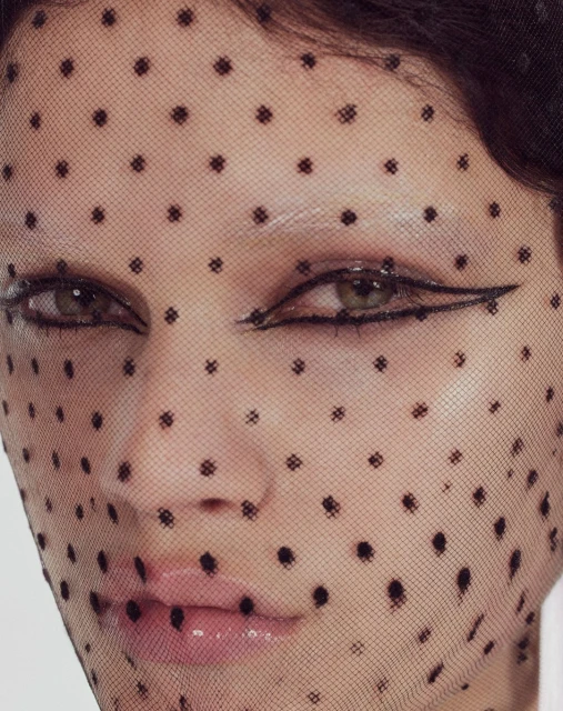 Close-up portrait of a woman with fair skin, visible makeup, and a dotted veil obscuring her face, set against a neutral backdrop.