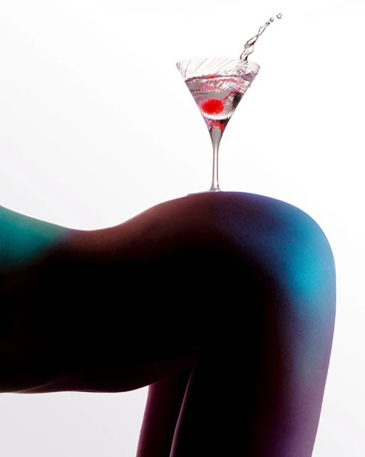 Martini glass with red cherry and splash of liquid balanced on the back of a posed figure with teal and purple gradient lighting.