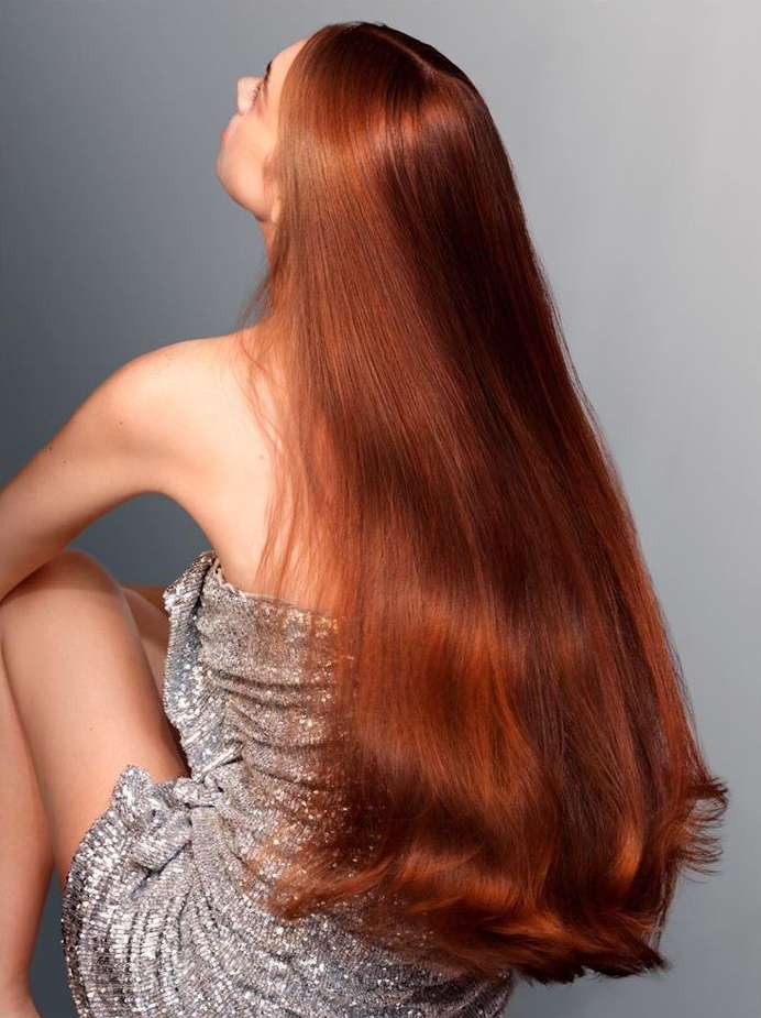From behind, a woman with long, copper hair wears a silver, sequined dress, sitting against a grey background.