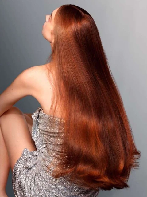 From behind, a woman with long, copper hair wears a silver, sequined dress, sitting against a grey background.
