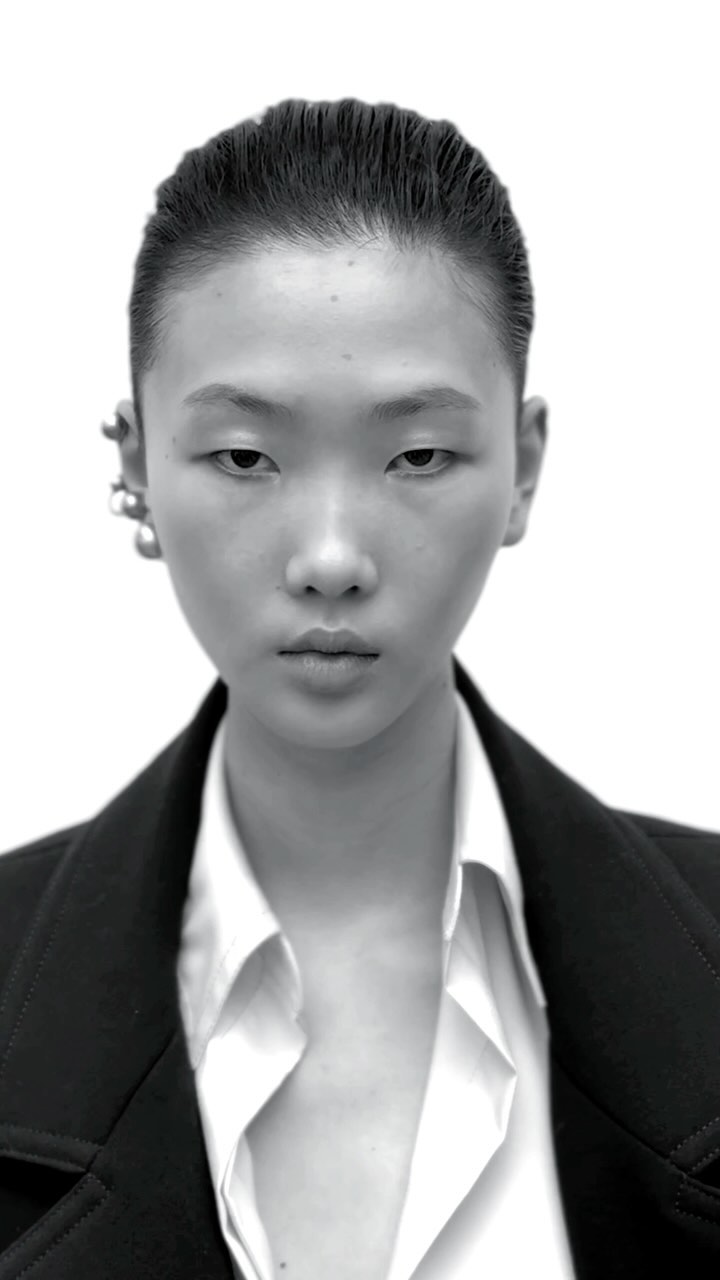 Close-up portrait of a model with slicked-back hair wearing a white collared shirt and black jacket against a plain white background