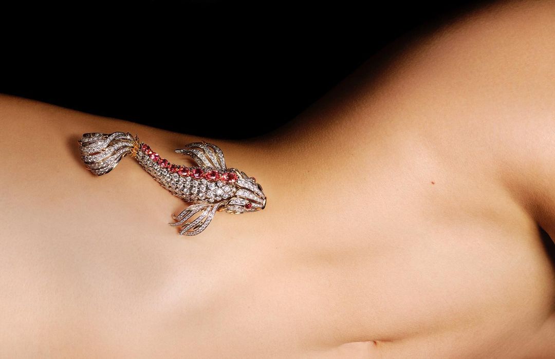 Close-up on the upper torso of a person with tanned skin featuring a gold and gemstone fish brooch jewelry on the upper chest.