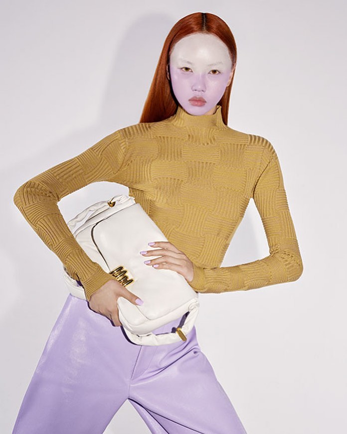 Fashionable woman with orange hair holds a white purse wearing a mustard-colored turtleneck and lilac pants in a studio shot.