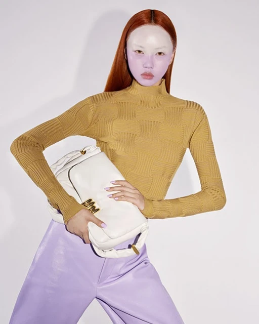 Fashionable woman with orange hair holds a white purse wearing a mustard-colored turtleneck and lilac pants in a studio shot.