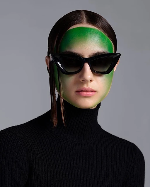 A woman with green makeup and black sunglasses poses in a studio with a gray background, wearing a black turtleneck sweater.