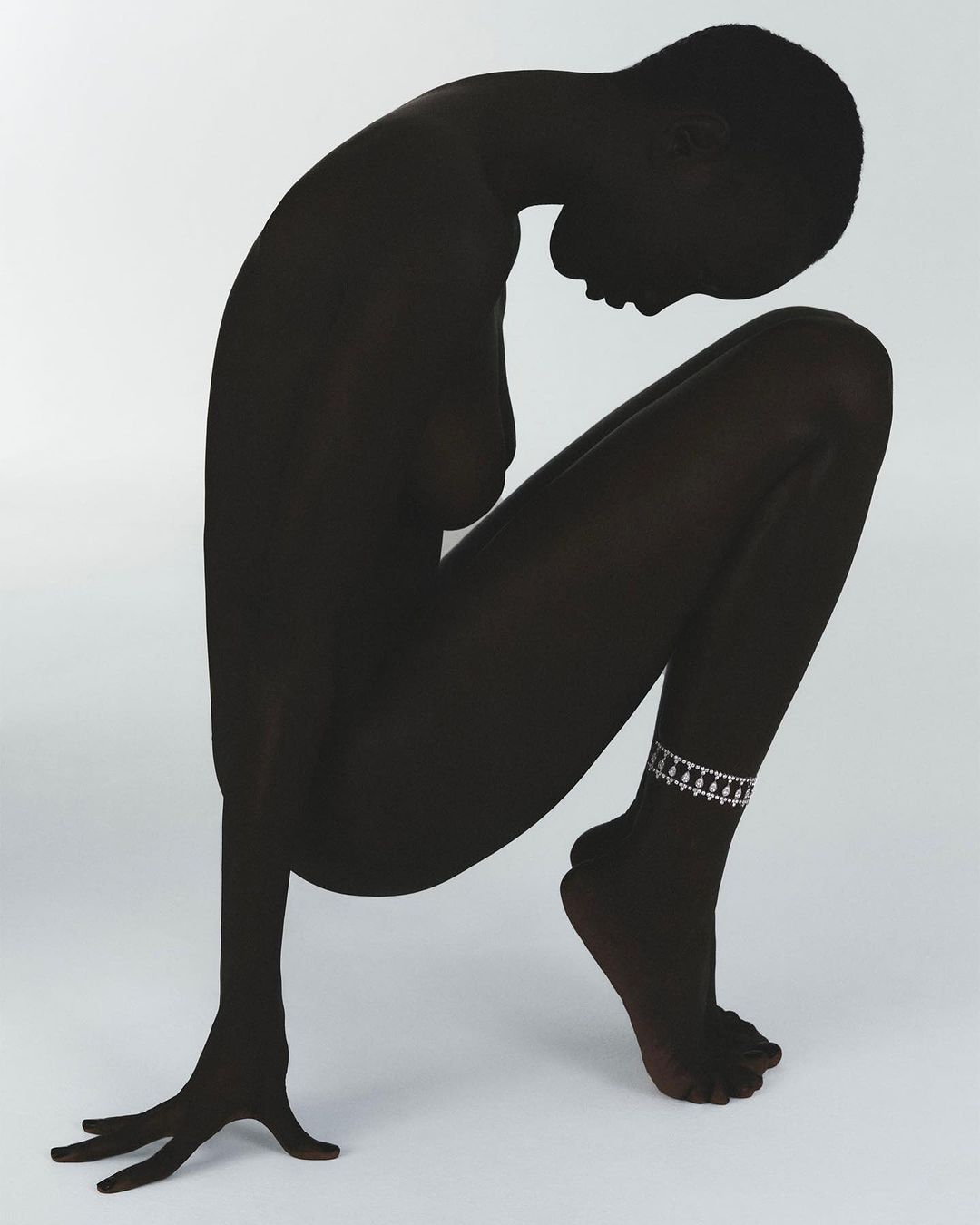 Silhouette of a Black woman crouching on a white backdrop wearing an anklet. Studio shot with a minimalist aesthetic.