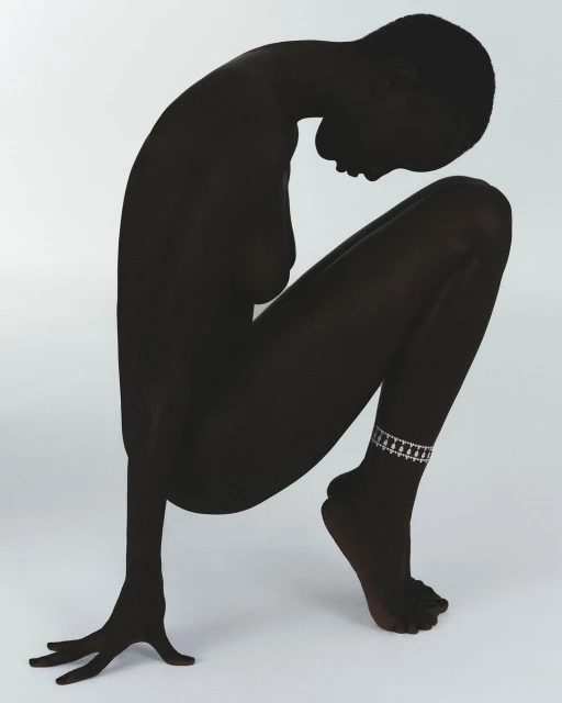 Silhouette of a Black woman crouching on a white backdrop wearing an anklet. Studio shot with a minimalist aesthetic.