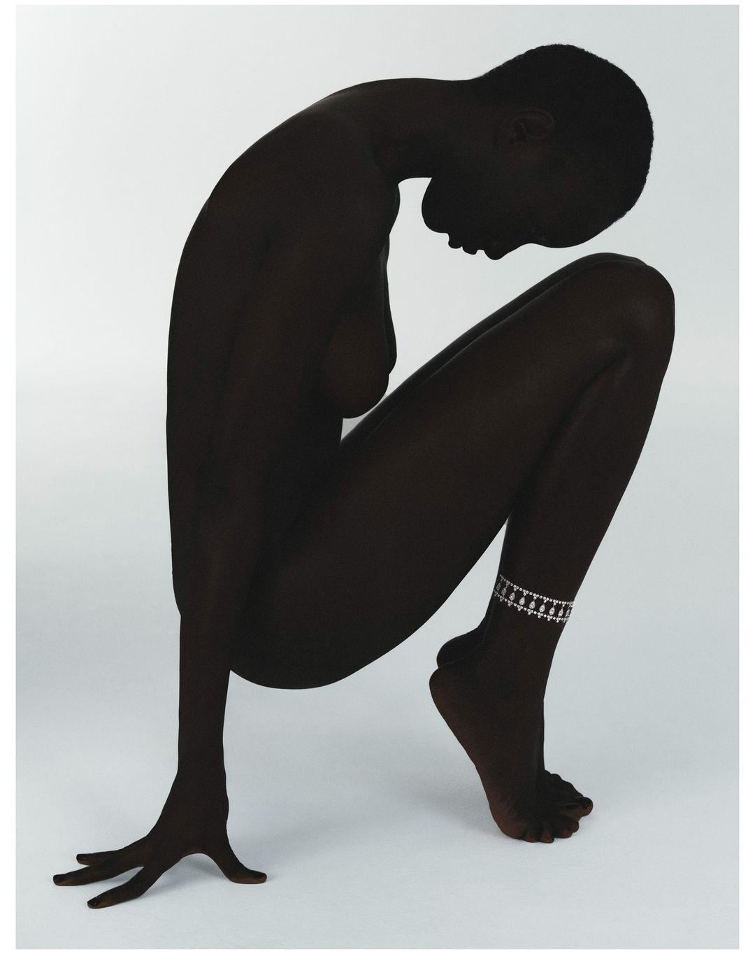 Artistic nude studio shot of a dark-skinned adult female in a crouched pose with head down and an anklet on her left ankle against a white background.